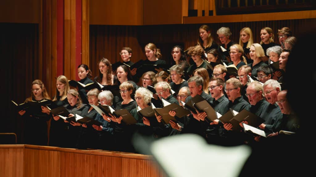 The Bach Choir sing passionately whilst looking at the conductor (who is off-screen) at the Southbank Centre's Royal Festival Hall. Another singer is at the front of the frame but is silhouetted and out of focus